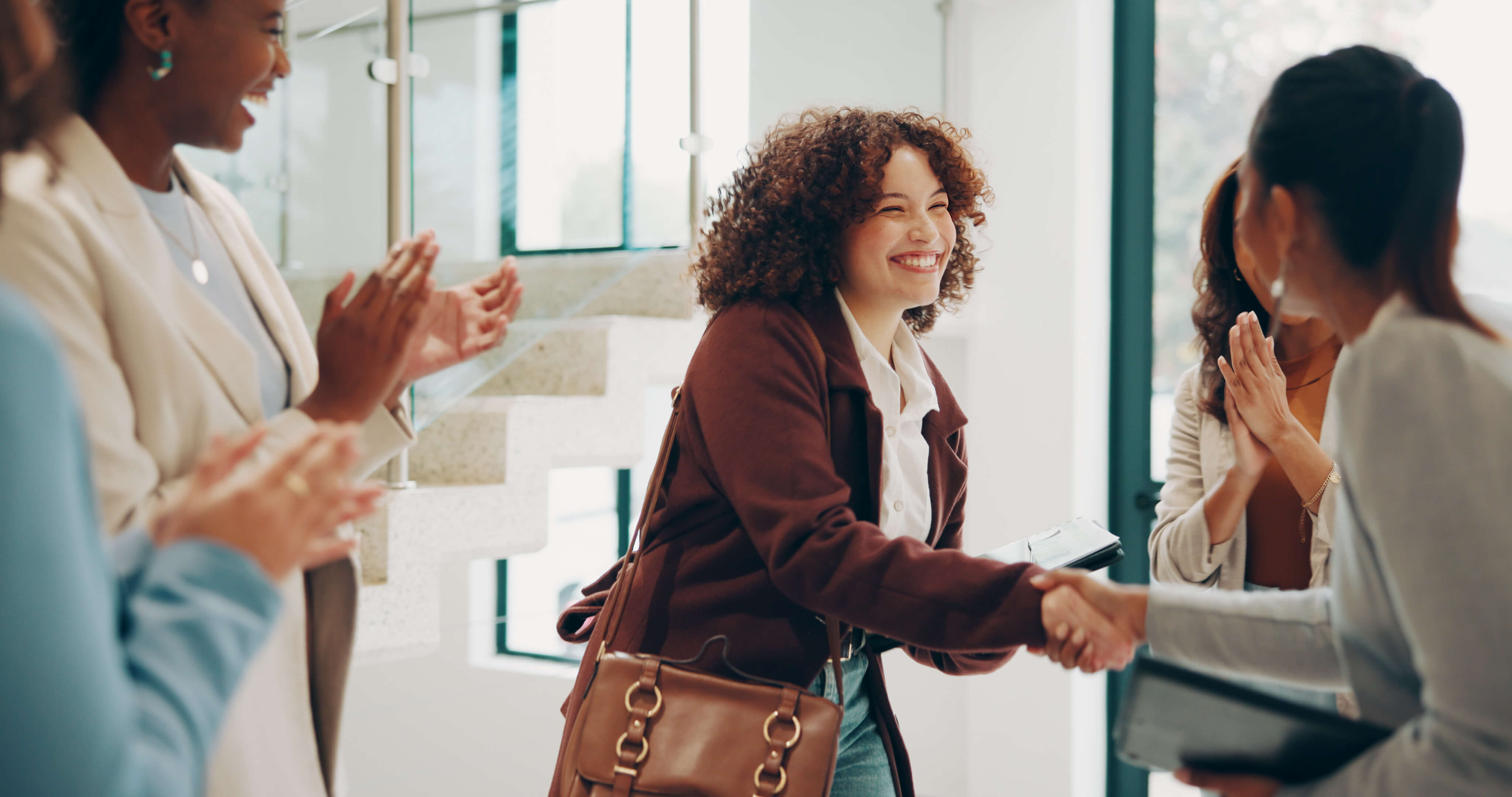 Woman in brown jacket and shoulder bags shakes hands with new members of her team, with others clapping around her